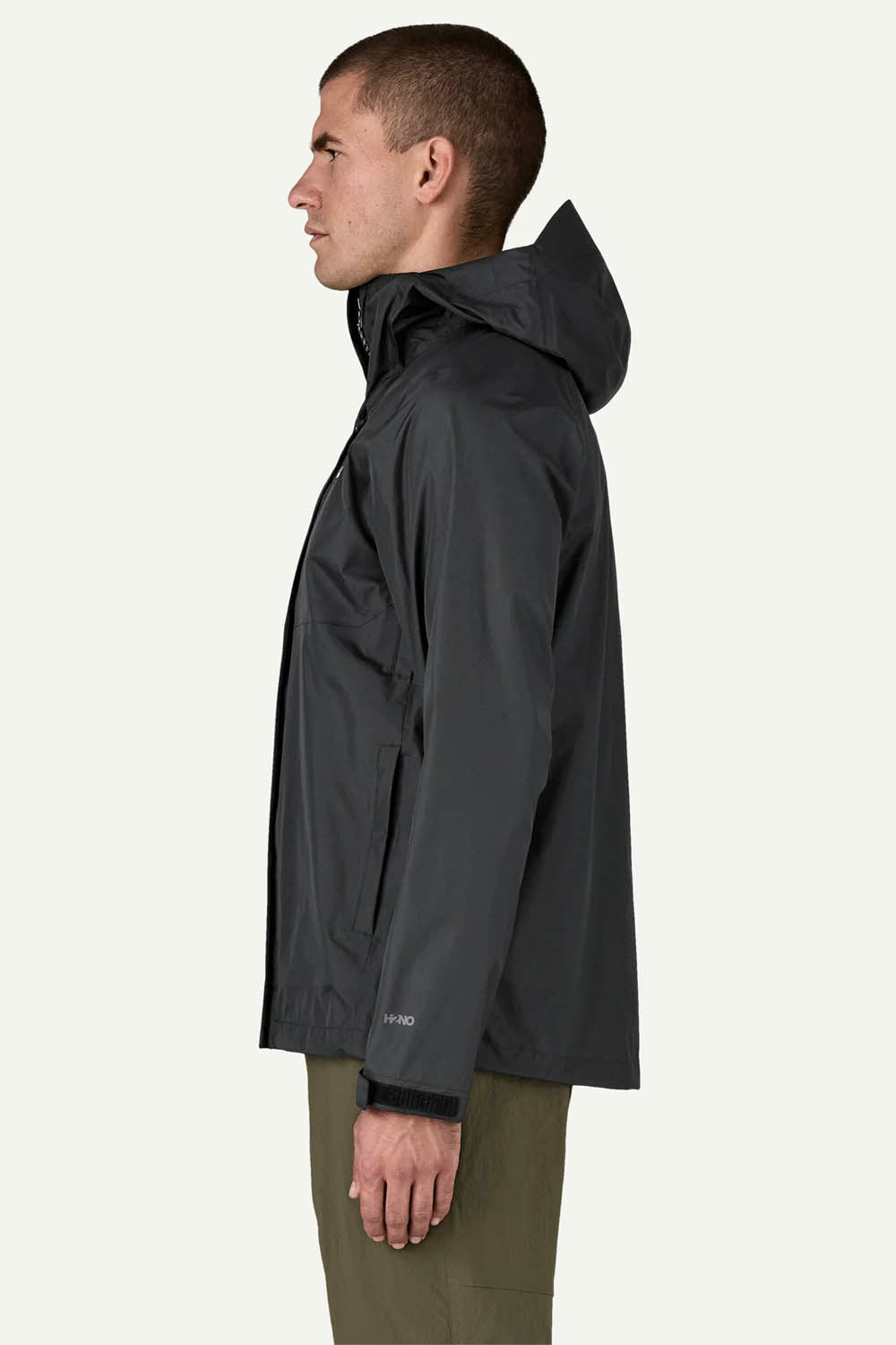 A man shown in profile wearing a dark gray hooded jacket and olive green pants, photographed against a plain light gray studio background.