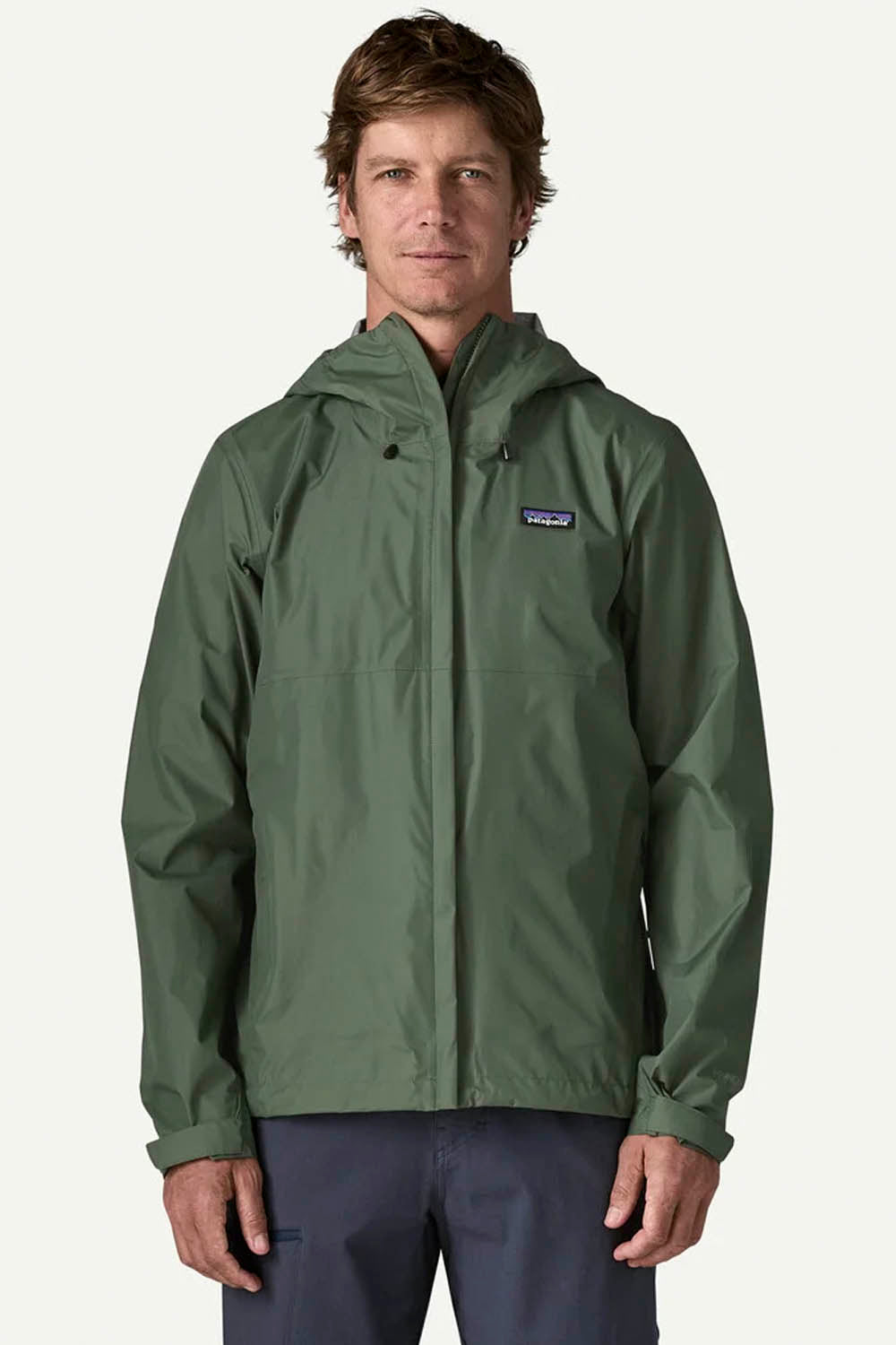 A man wearing a green Patagonia hooded rain jacket and navy pants, standing against a light off-white background.