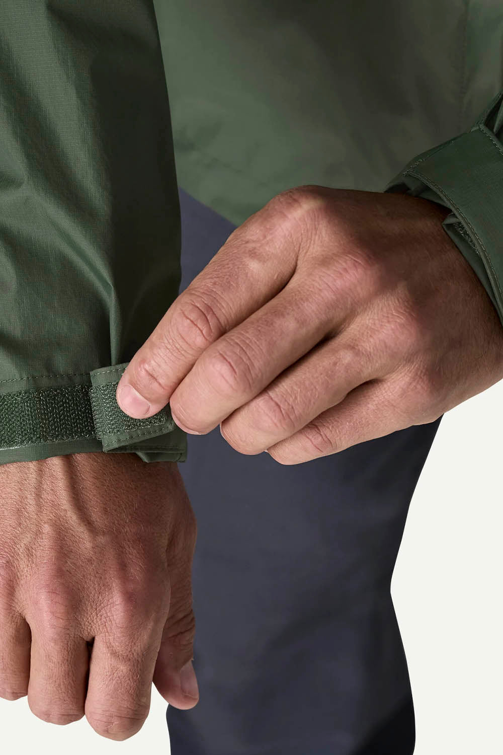 Close-up of a person's hands adjusting the cuff of an olive green waterproof jacket, with dark navy pants visible underneath against a light off-white background.