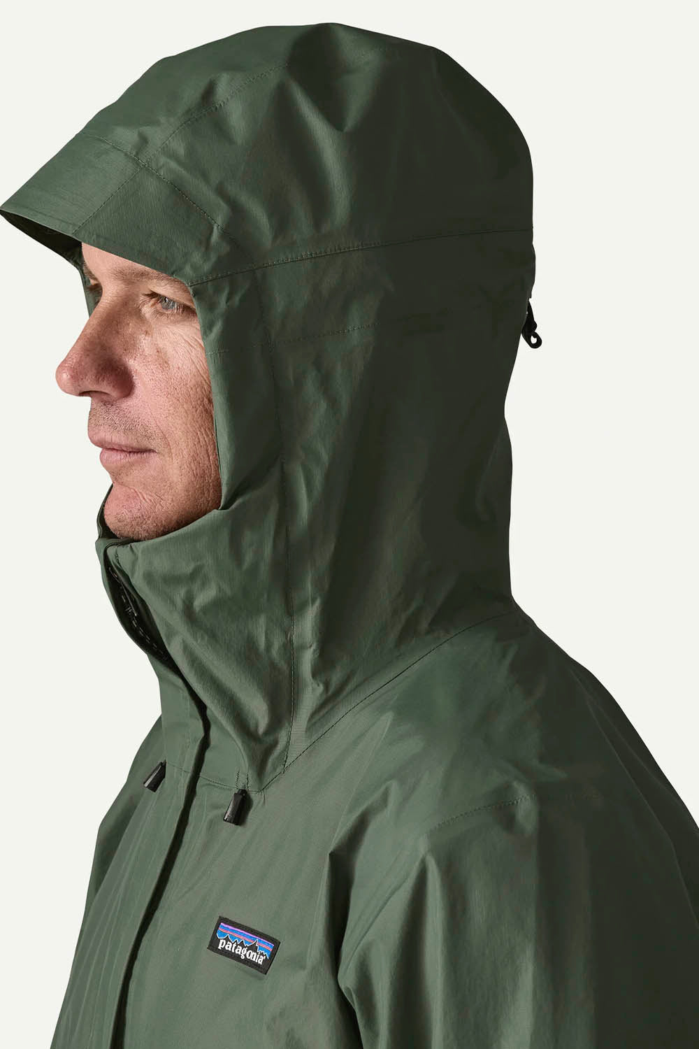 A man wearing a Patagonia hooded rain jacket in olive green, shown in profile against a light off-white background. The Patagonia logo is visible on the chest.