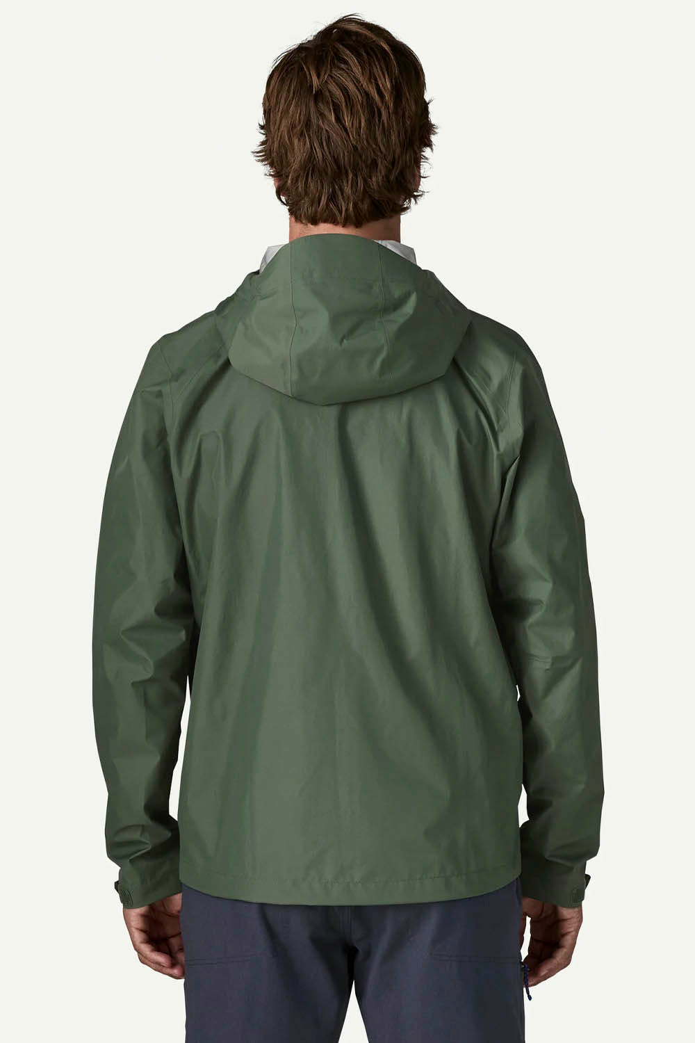 A man viewed from behind wearing an olive green hooded jacket and navy pants against a light off-white background.