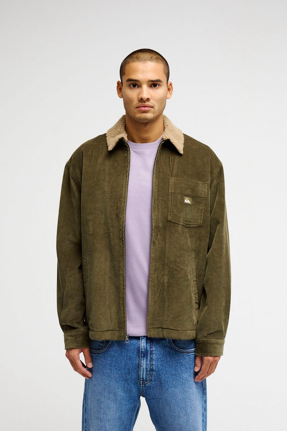 A man wearing an olive green corduroy jacket with a beige sherpa collar, layered over a light purple t-shirt and blue jeans, standing against a light gray studio background.