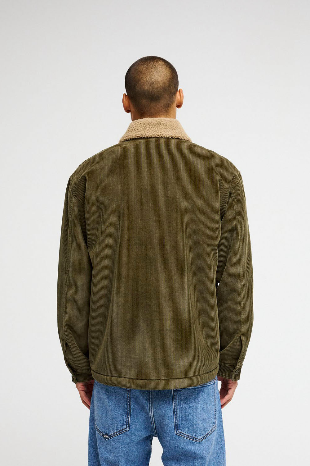 A man viewed from behind wearing an olive green corduroy jacket with a beige sherpa collar and blue jeans, against a plain light gray studio background.