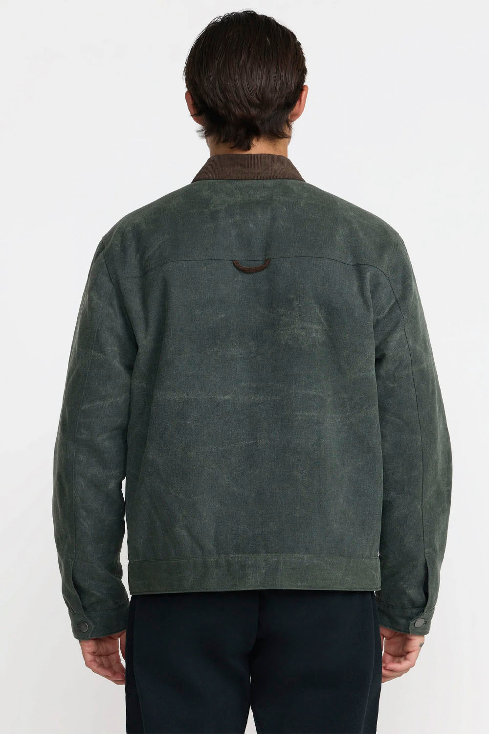 A man wearing a dark green waxed canvas utility jacket with a brown corduroy collar, standing against a plain white studio background.