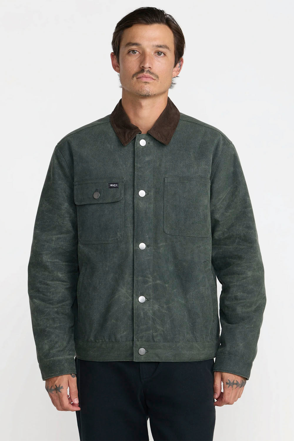 A man wearing a dark green waxed canvas utility jacket with a brown corduroy collar, standing against a plain white studio background.