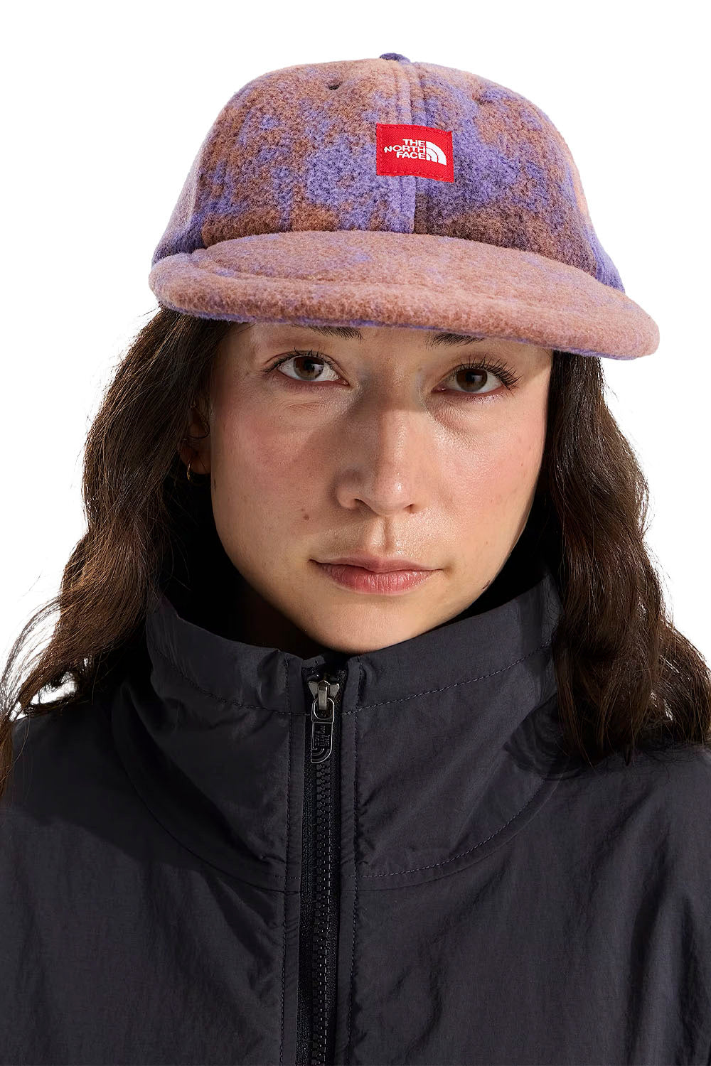 A woman wearing a textured, heathered purple and brown baseball cap with a red The North Face logo patch, paired with a dark gray high-collar jacket, against a plain white background.