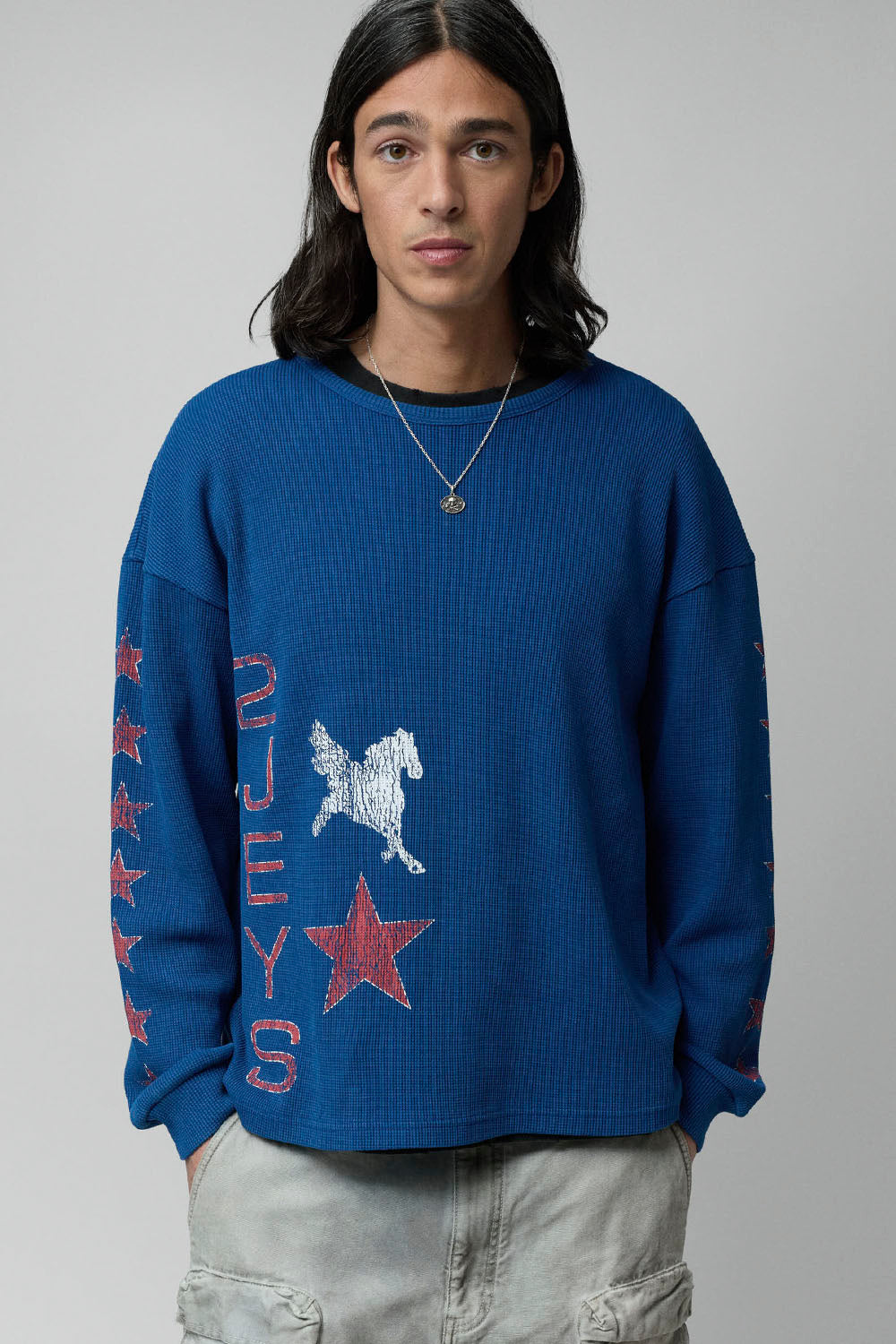 A model wearing a royal blue waffle-knit crewneck sweatshirt with red and white graphic prints, paired with light gray cargo pants, against a neutral gray background.