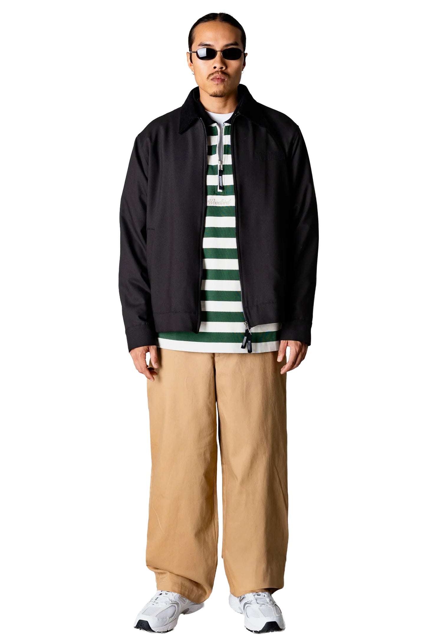 Person wearing a black Woodbird Wbpon Guan Work jacket, striped shirt, beige pants, and white sneakers on a white background