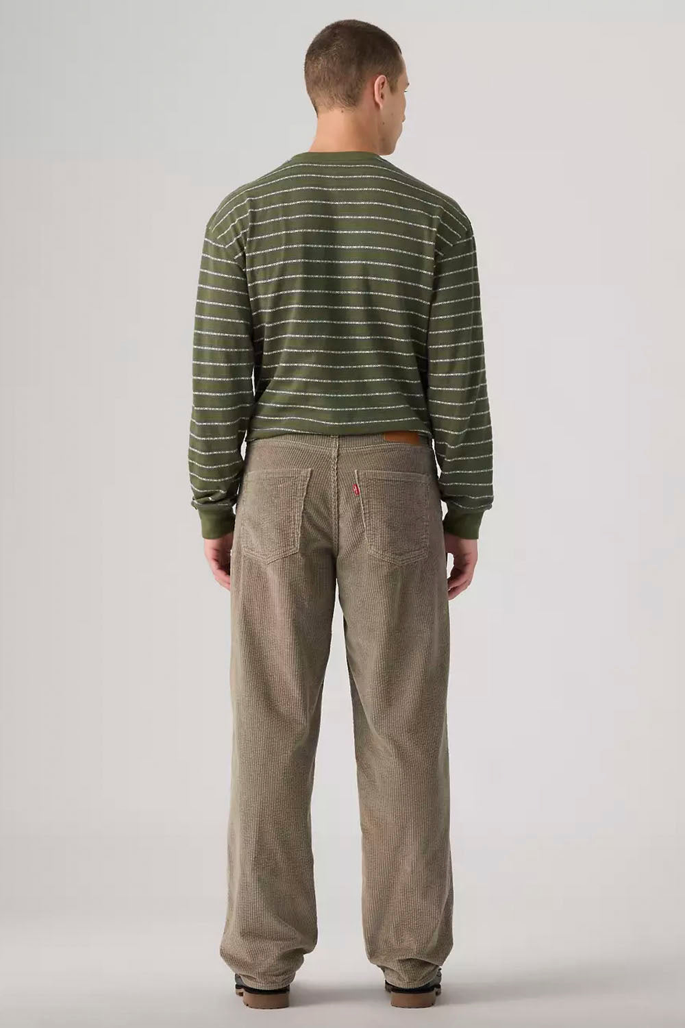 A man viewed from behind wearing a green and white striped long-sleeve shirt and beige corduroy pants, standing against a light gray studio background.