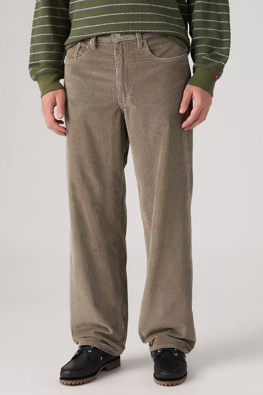 A person wearing loose-fit corduroy pants in a taupe-brown color, styled with a green striped sweater and black shoes, photographed against a light gray studio background.