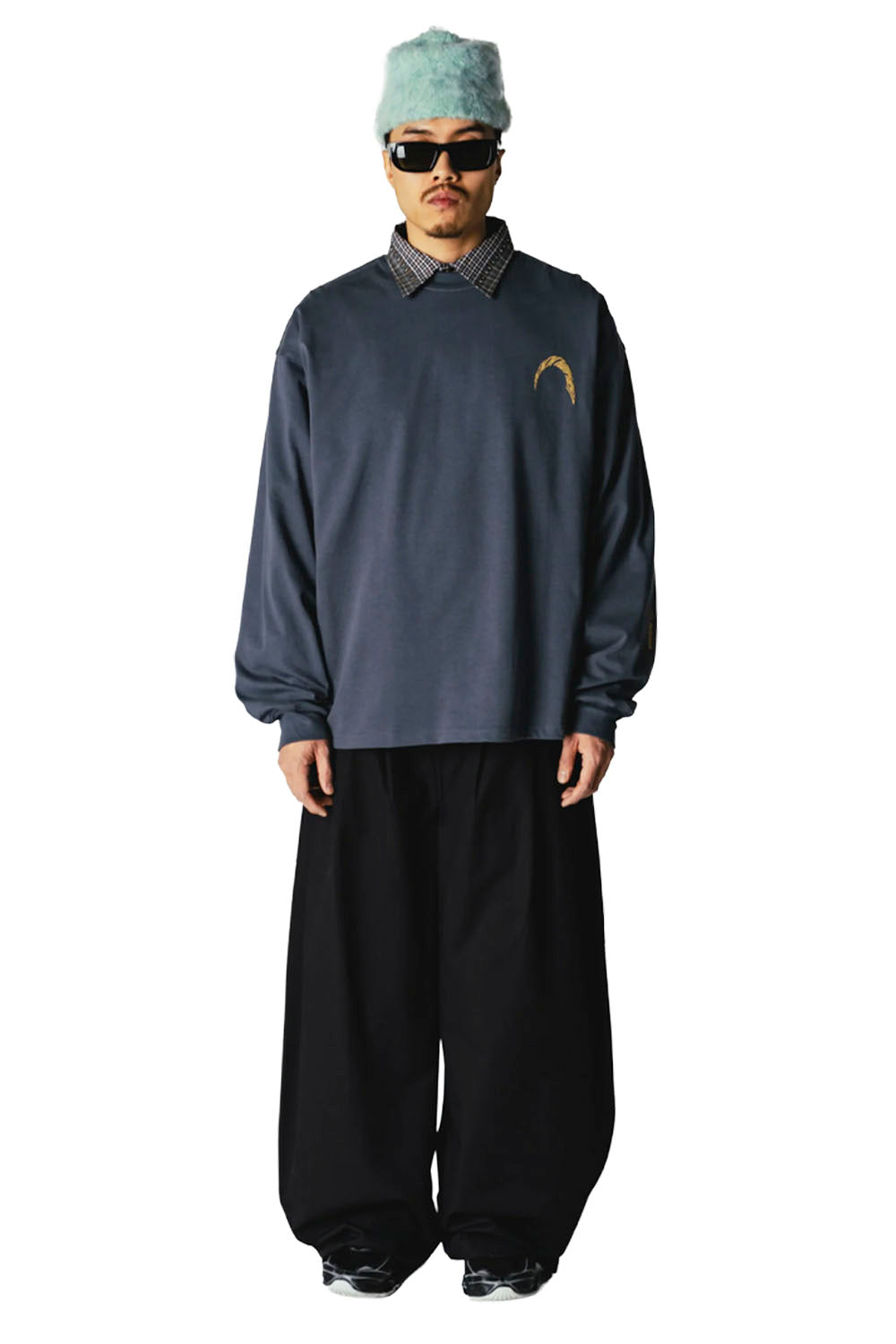 A model wearing a dark navy blue long-sleeve sweatshirt with a small gold graphic on the left chest, styled with black wide-leg pants, a patterned collared shirt, sunglasses, and a light blue fuzzy hat against a white background.