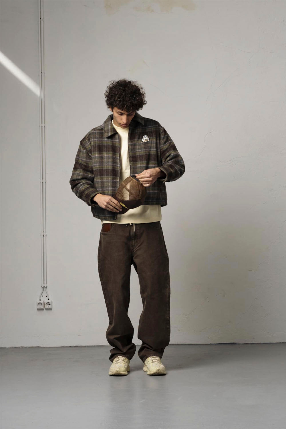 A man stands in a minimalist studio setting wearing a brown and black plaid overshirt over a cream t-shirt, dark brown pants, and off-white sneakers. He is holding a brown cap in his hands.