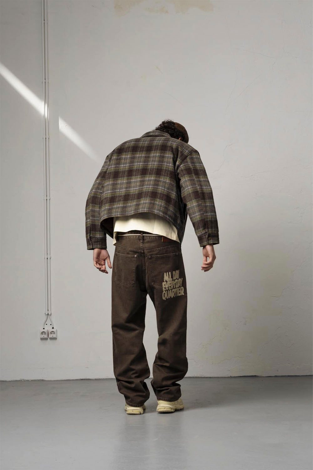 A person viewed from behind wearing a brown and beige plaid jacket, a white undershirt, and dark brown pants with 'ALL DOGS ESTABLISH QUARTER' printed in gold on the back pocket, standing in a minimalist studio with a white wall and gray floor.