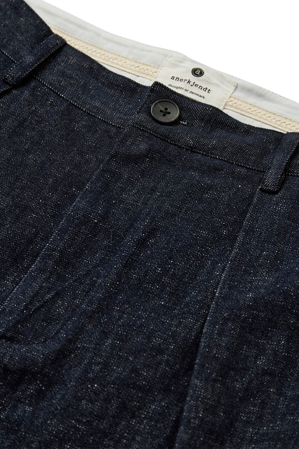 Close-up of dark indigo denim fabric showing the texture, a button closure, and a cream-colored inner waistband label with the brand name 'ashford'.