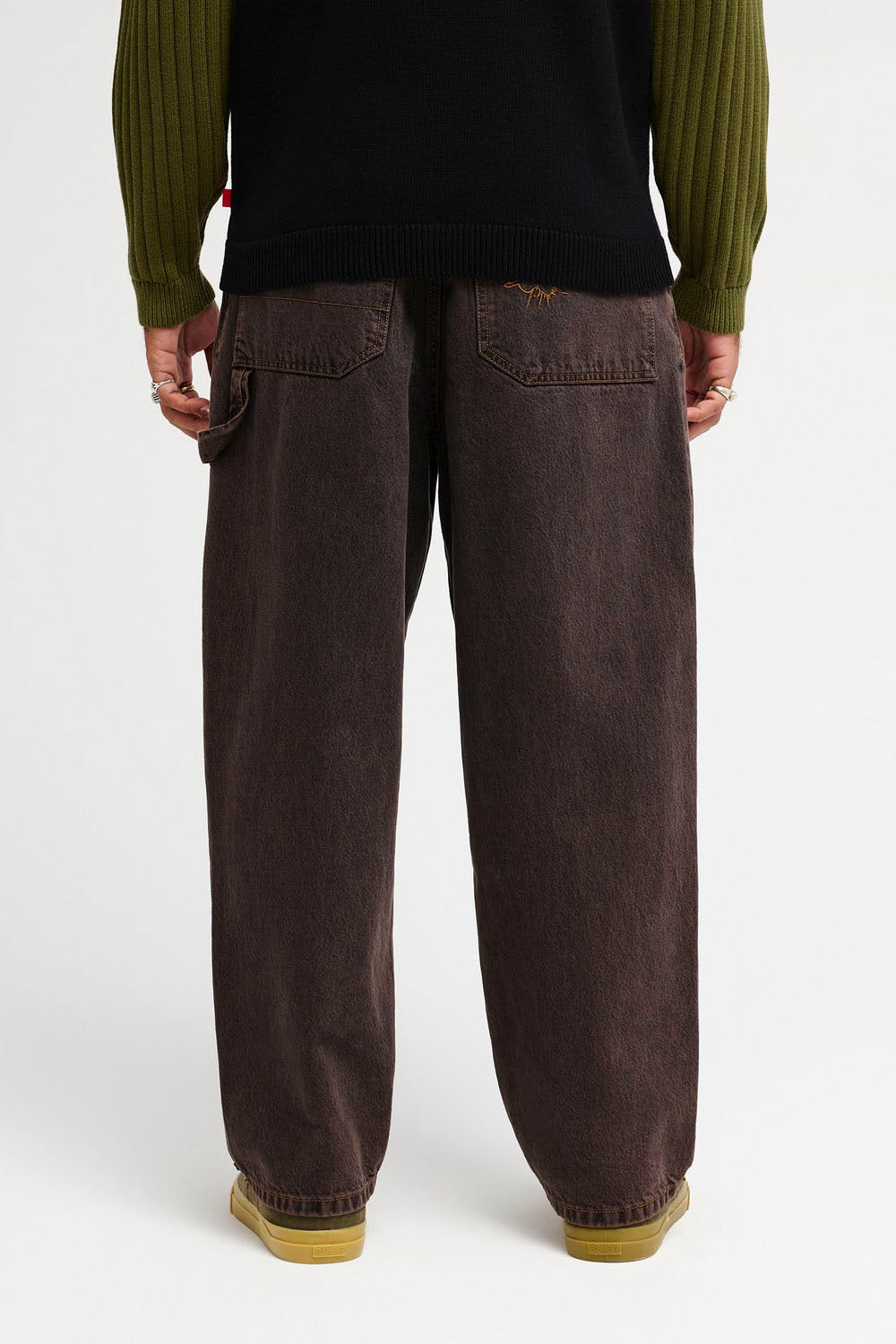 A person shown from the back wearing very wide-leg dark brown washed denim jeans, a black ribbed sweater, and olive green long-sleeve shirt, paired with tan shoes, against a plain white studio background.