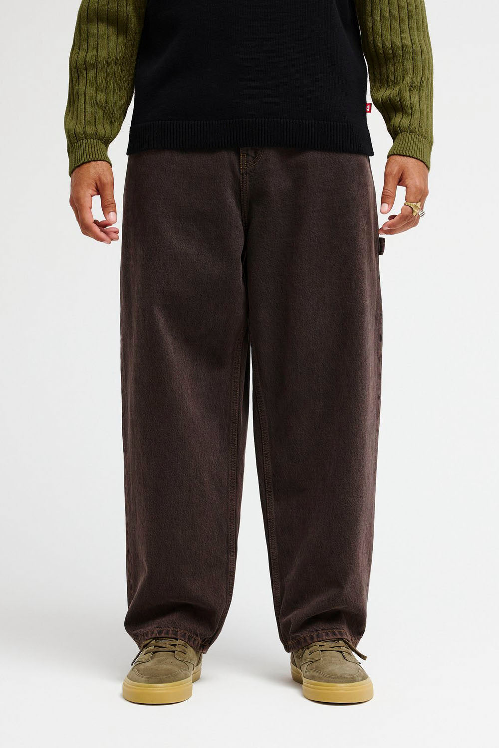 A person wearing loose dark brown corduroy pants, a black top, and olive green and tan sneakers, shown from the chest down against a plain light background.
