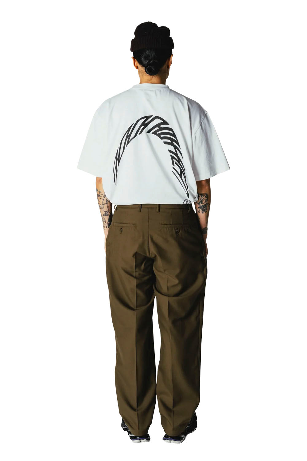 A person shown from the back wearing a white short-sleeve T-shirt with a large black curved graphic on the back, paired with dark olive brown pants, against a plain white background.