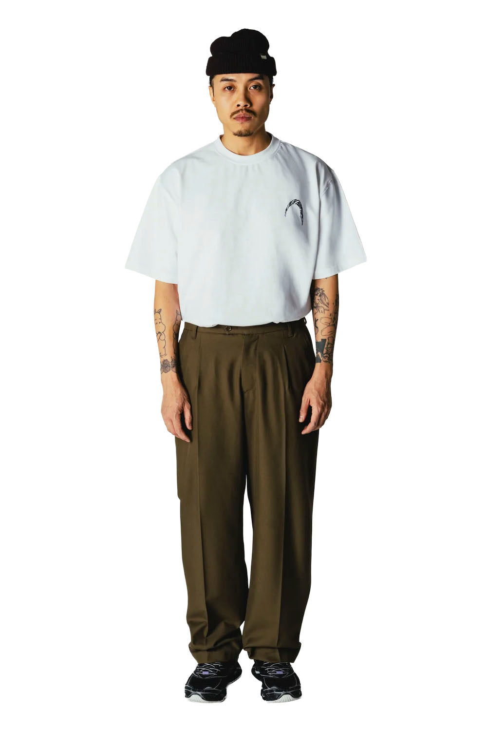 A man stands against a white studio background wearing a light blue oversized short-sleeve T-shirt with a small chest graphic, loose olive-brown pants, black sneakers, and a black beanie.