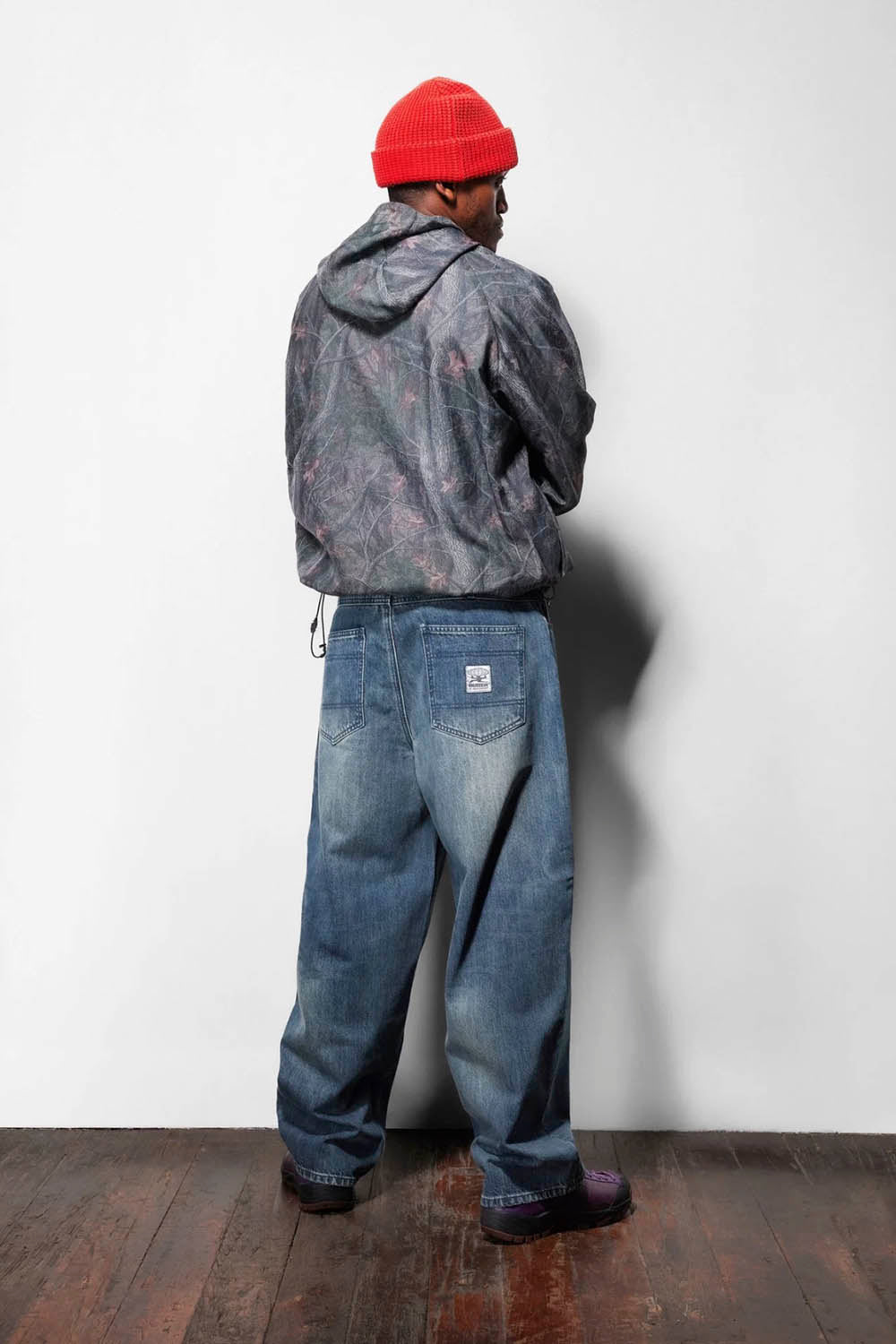 A person standing with their back to the camera, wearing a red knit beanie, a dark gray hooded jacket, and wide-leg blue jeans, set against a plain white wall and dark wooden floor.