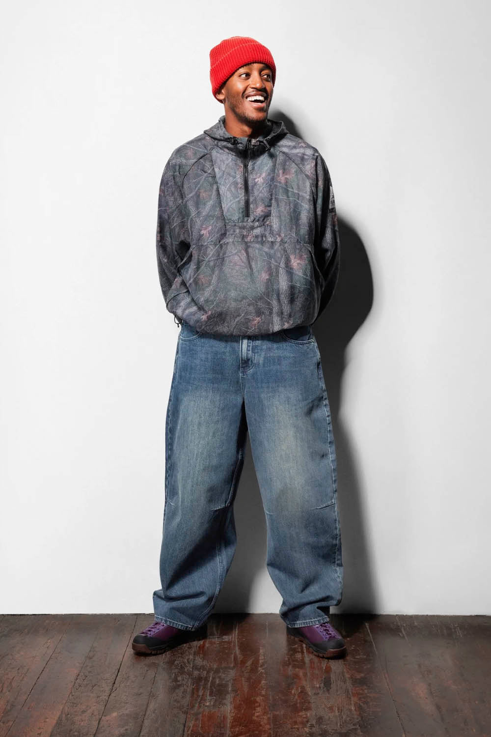 A man wearing a red beanie, a dark gray patterned hooded sweatshirt, baggy blue jeans, and purple sneakers, standing against a white wall on a dark wood floor.