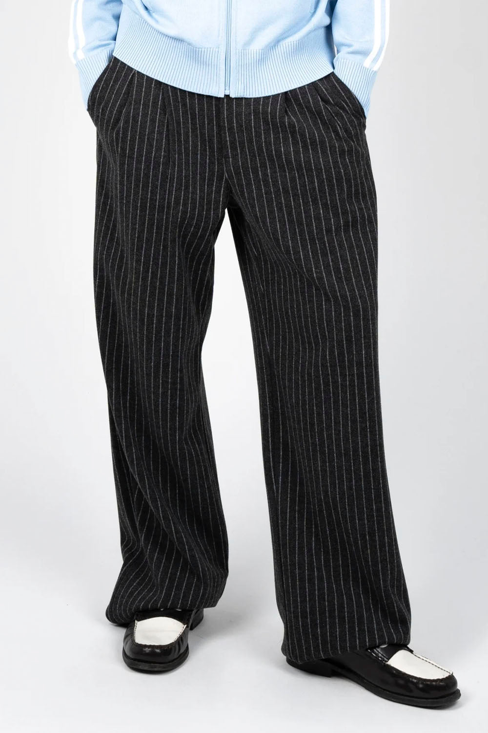 Black pinstripe wide-leg trousers worn by a model, styled with a light blue zip-up jacket and black-and-white loafers, shot against a light gray studio background.