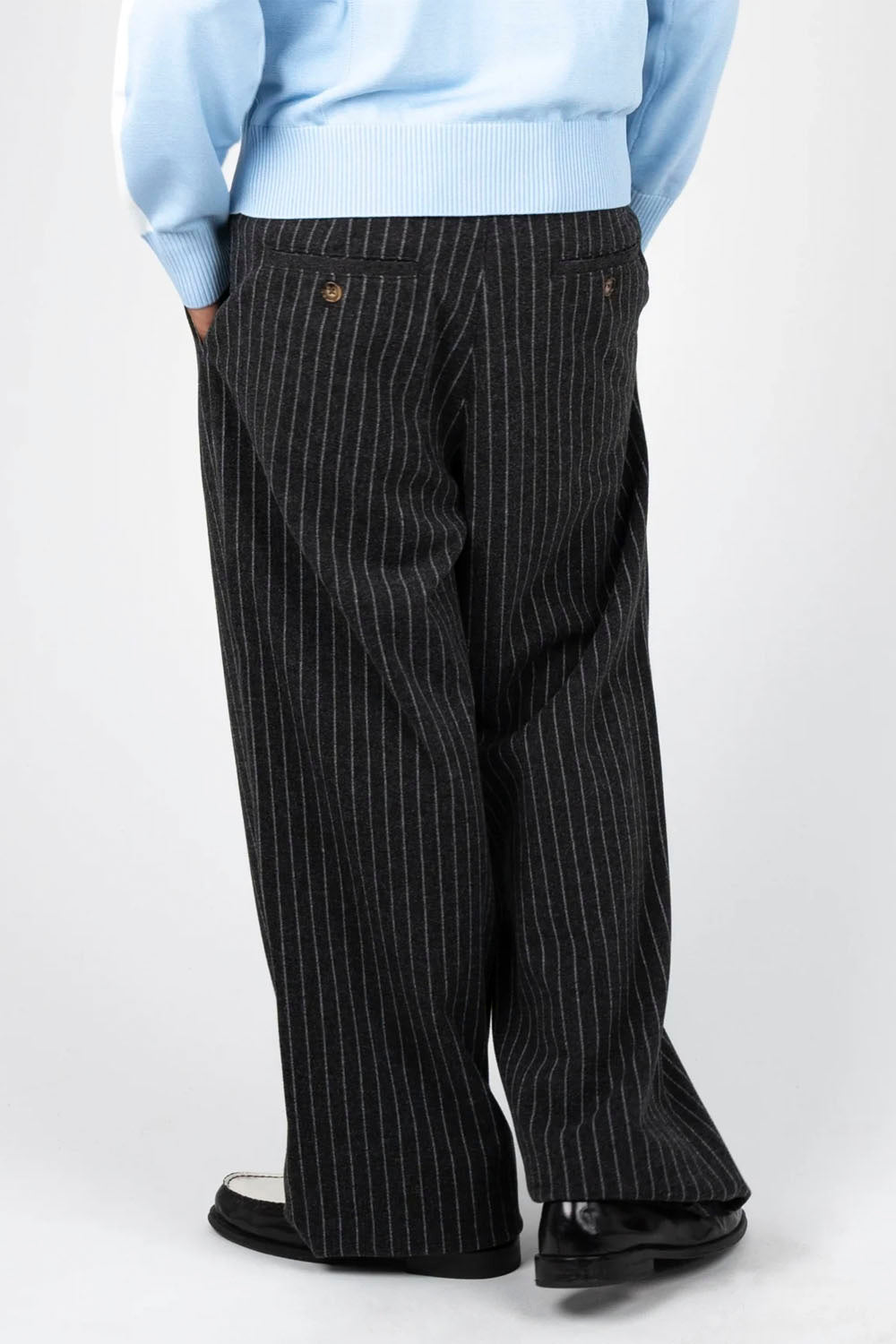 Black pinstripe wide-leg trousers worn by a model, styled with a light blue zip-up jacket and black-and-white loafers, shot against a light gray studio background.