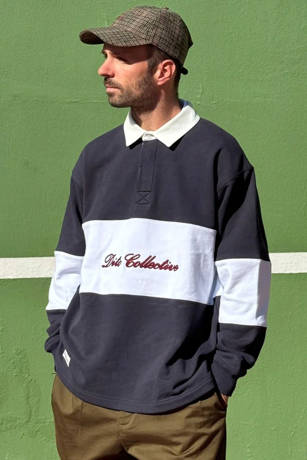 A man wearing a dark navy and white color-blocked rugby pullover with 'Art Collective' embroidered on the chest, paired with a brown plaid cap and khaki pants, standing against a green wall.