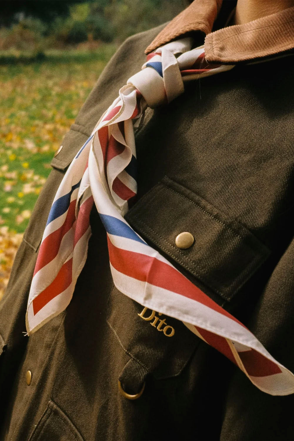 A close-up lifestyle shot of a person wearing a brown corduroy jacket with a striped silk scarf in red, white, and blue tied around the collar. The jacket has gold buttons and a gold embroidered 'Dito' logo on the chest pocket.