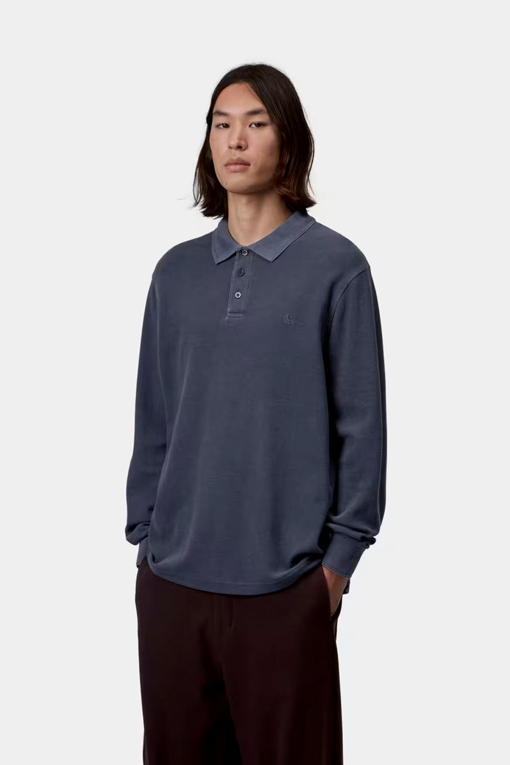 A model stands against a light gray studio background wearing a muted slate blue long-sleeve polo shirt and dark brown wide-leg trousers.