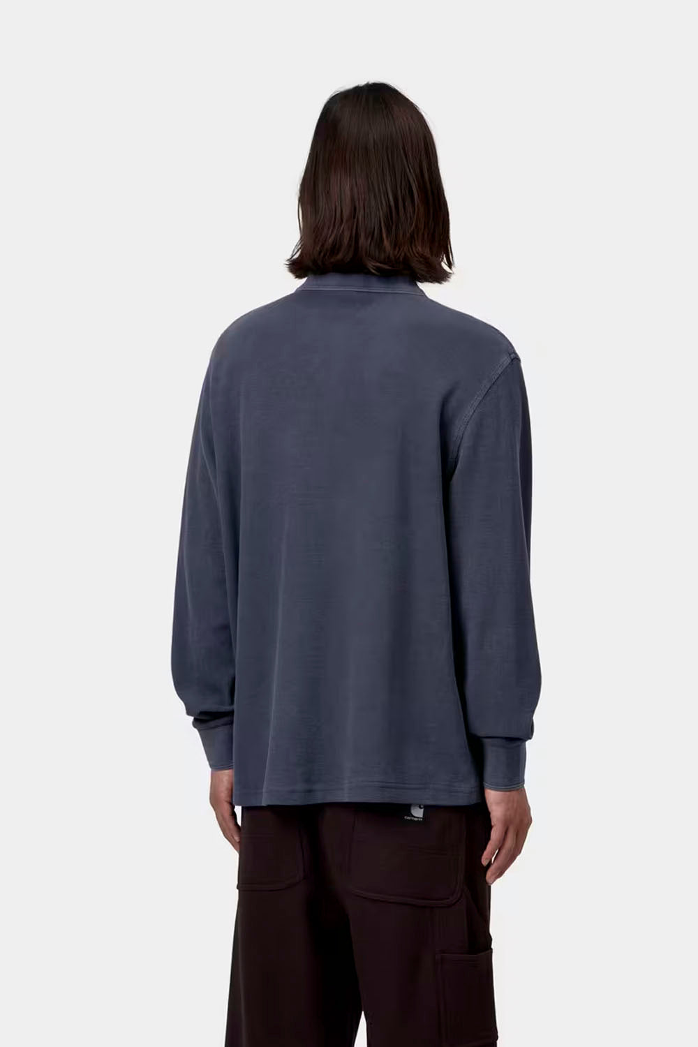 A model stands against a light gray studio background wearing a muted slate blue long-sleeve polo shirt and dark brown wide-leg trousers.