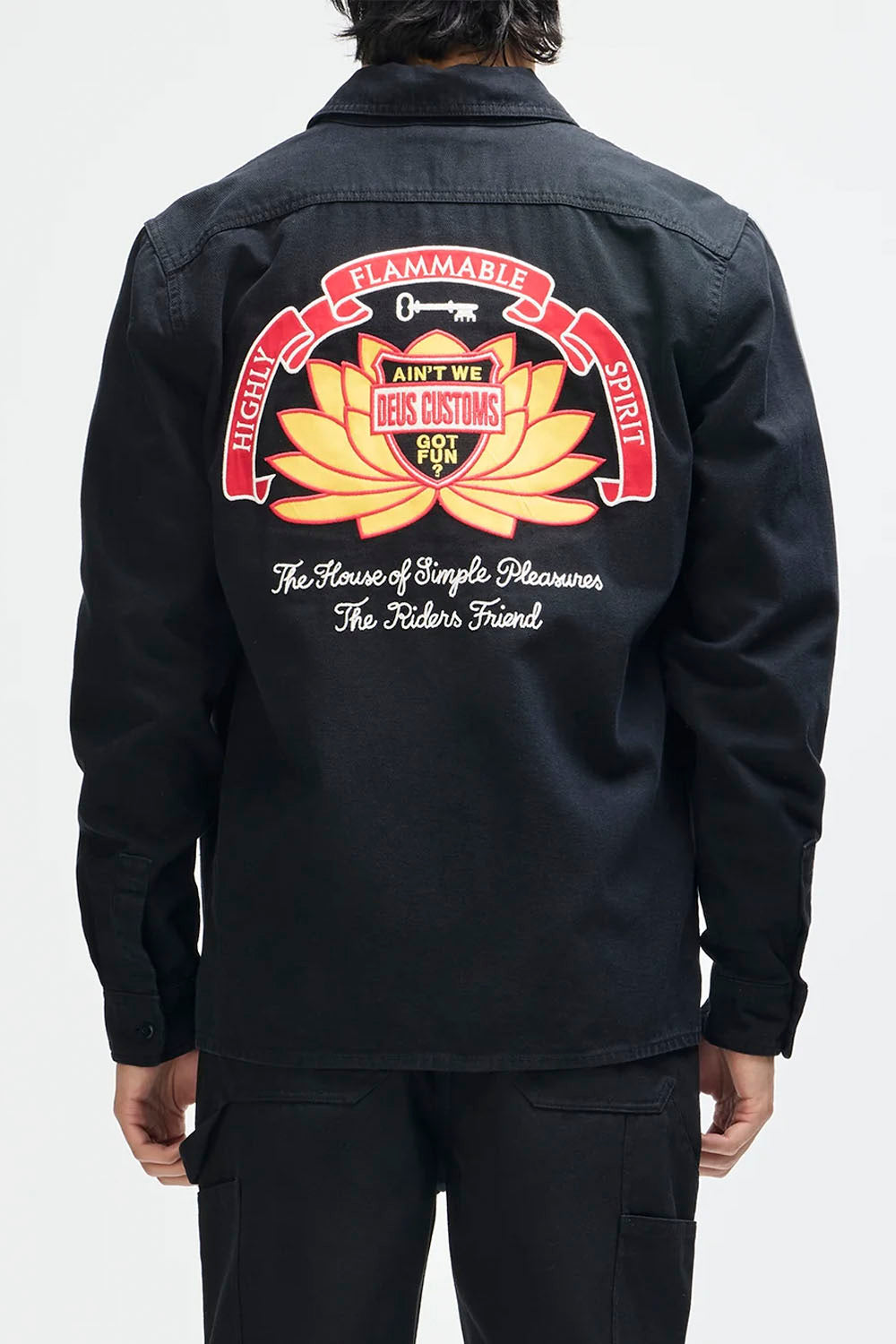 A person shown from the back wearing a black overshirt with a large colorful back graphic. The graphic features red banners, a yellow-orange lotus flower, and white script text reading 'The House of Simple Pleasures The Rider's Friend'.