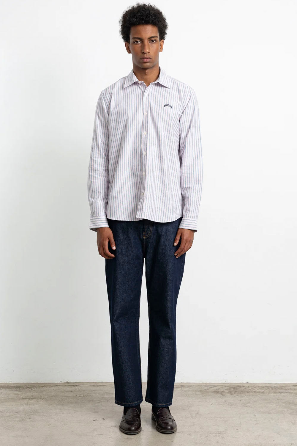 A man wearing a white long-sleeve button-down shirt with thin vertical blue stripes, paired with dark blue jeans and dark brown leather shoes, standing against a plain white wall.