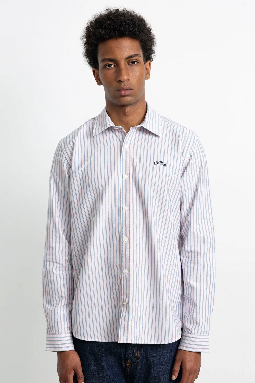 A man wearing a white long-sleeve button-down shirt with thin vertical navy blue stripes and a small embroidered logo on the left chest, paired with dark blue jeans, against a plain white studio background.