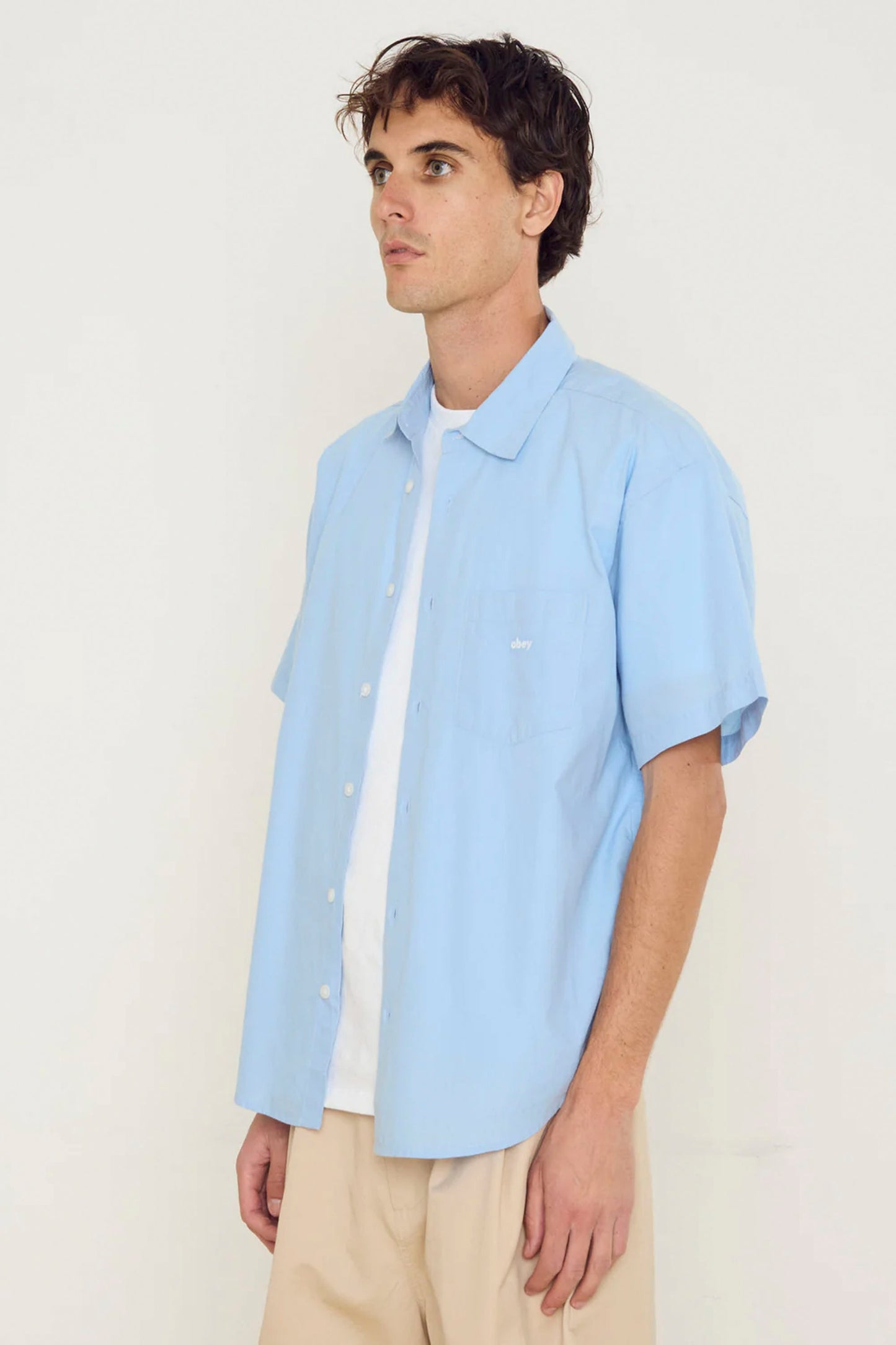 Man wearing a Light blue OBEY Bigwig Jamie shirt over a white t-shirt on a plain background