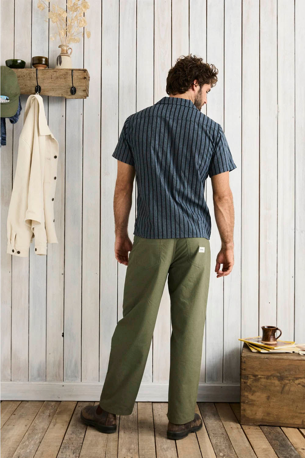 Man wearing a striped shirt and green pants standing in a room with wooden floor and white paneled walls.