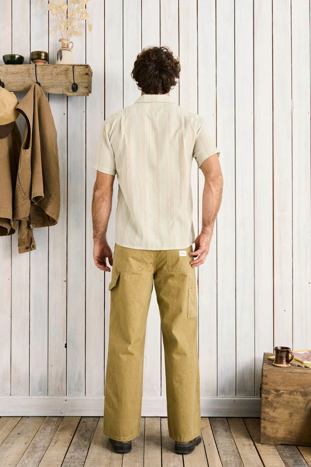 Man wearing a beige shirt and green pants standing in a room with wooden walls and floor.
