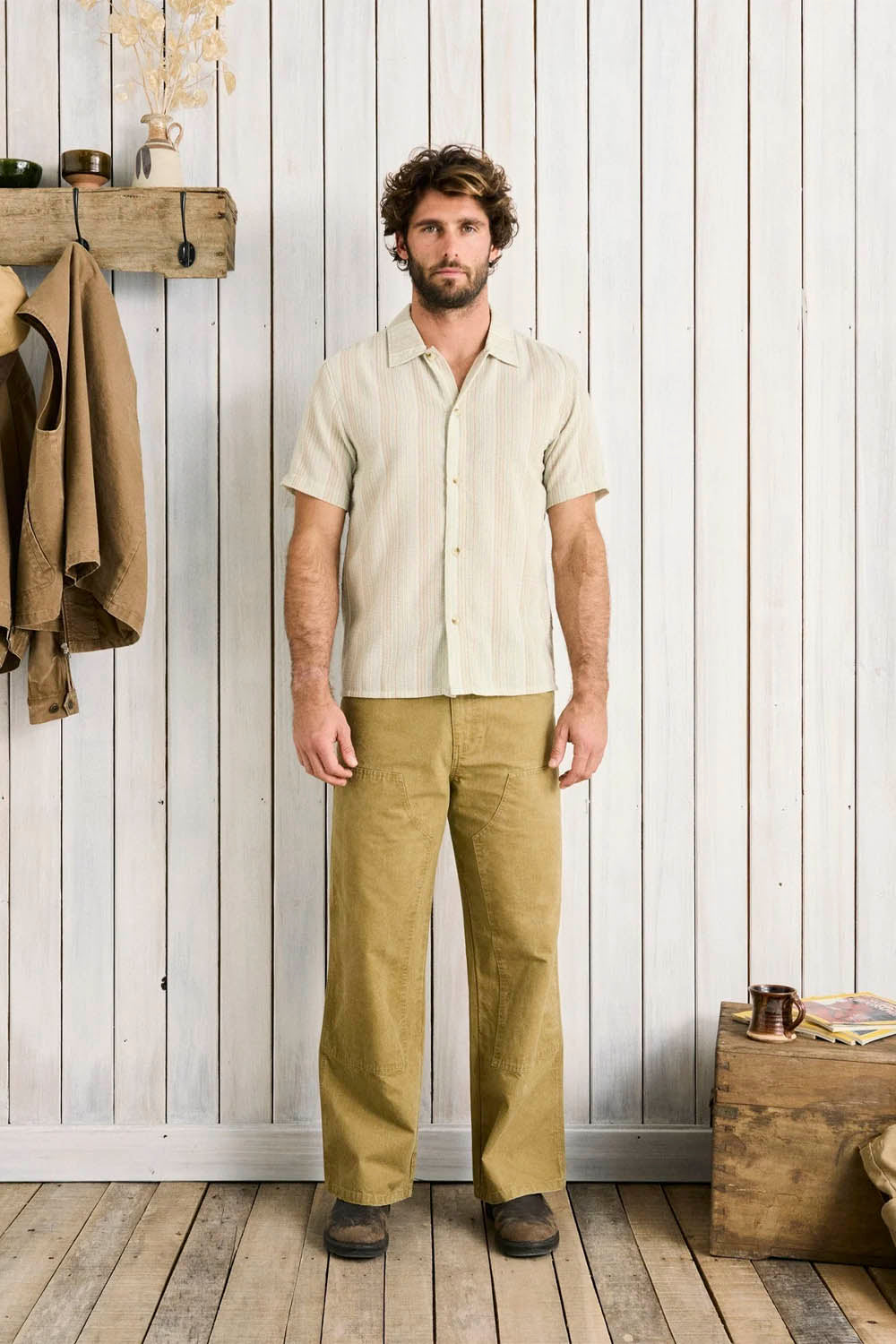 Man wearing a light-colored shirt and beige pants standing against a white paneled wall.