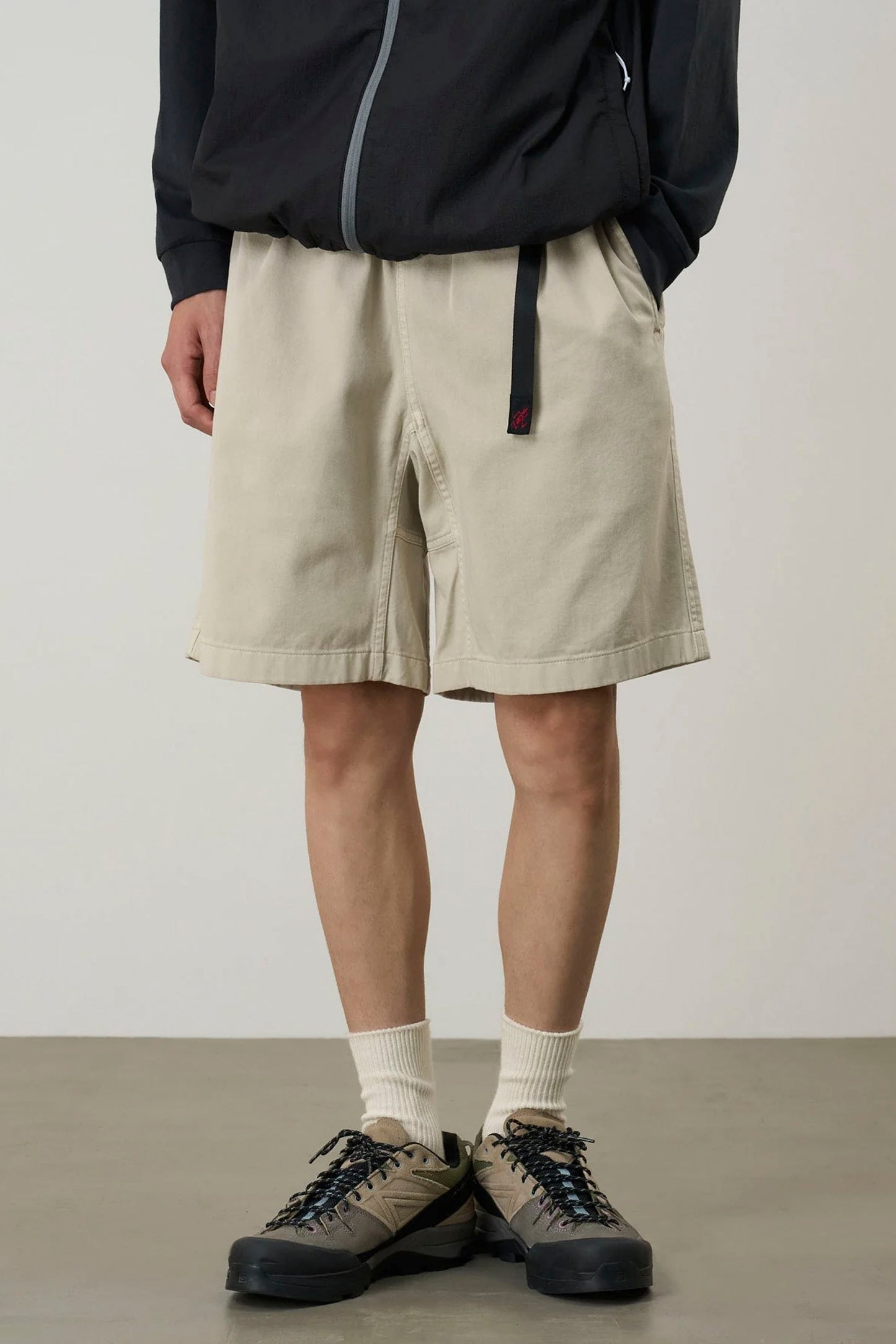 Person wearing beige Gramicci G-Short pigment shorts, black jacket, and hiking shoes on a plain background