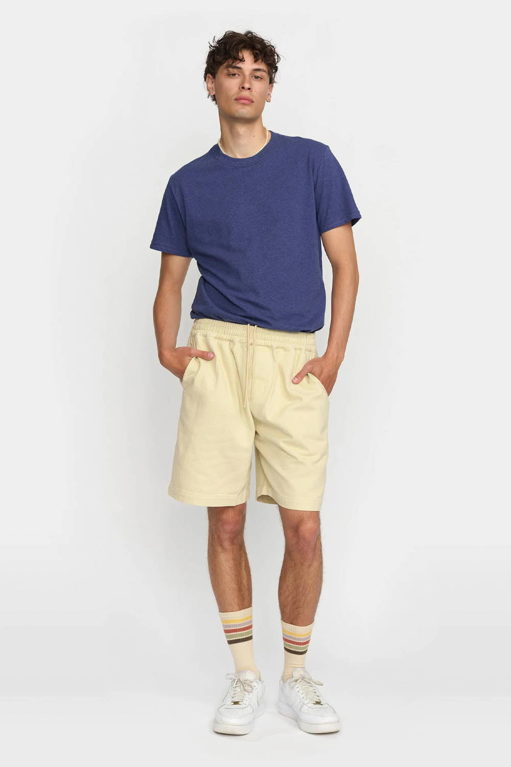 Man wearing a blue t-shirt, beige shorts, and white sneakers on a white background