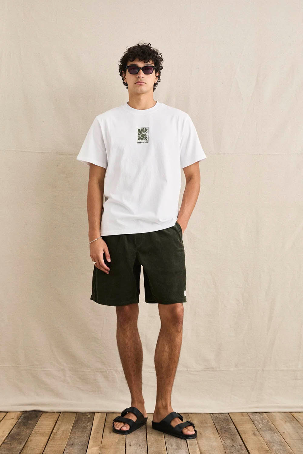 Man wearing a white t-shirt with a logo and dark shorts against a beige wall.