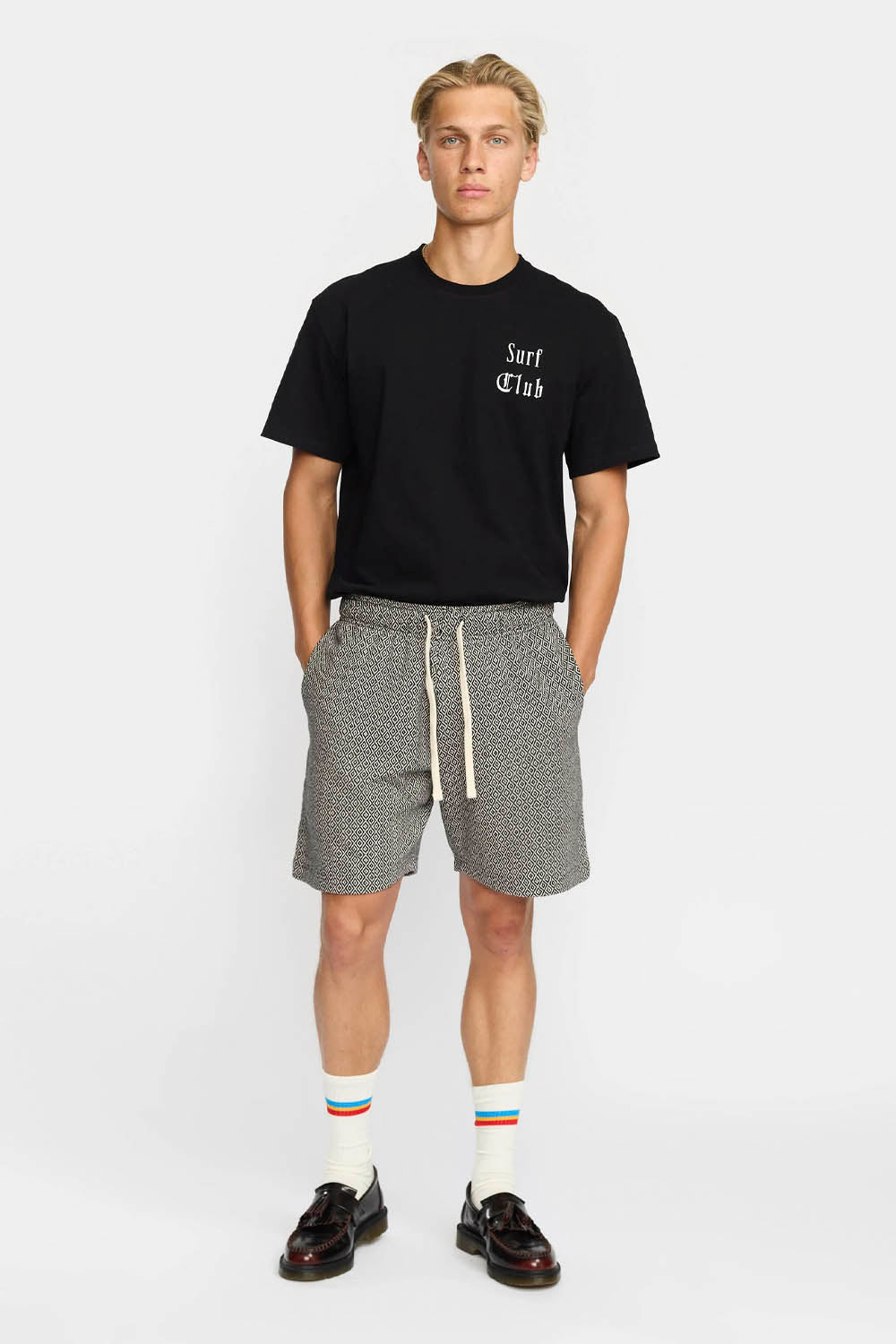 Man wearing a black t-shirt with 'Surf Club' text and patterned shorts on a white background