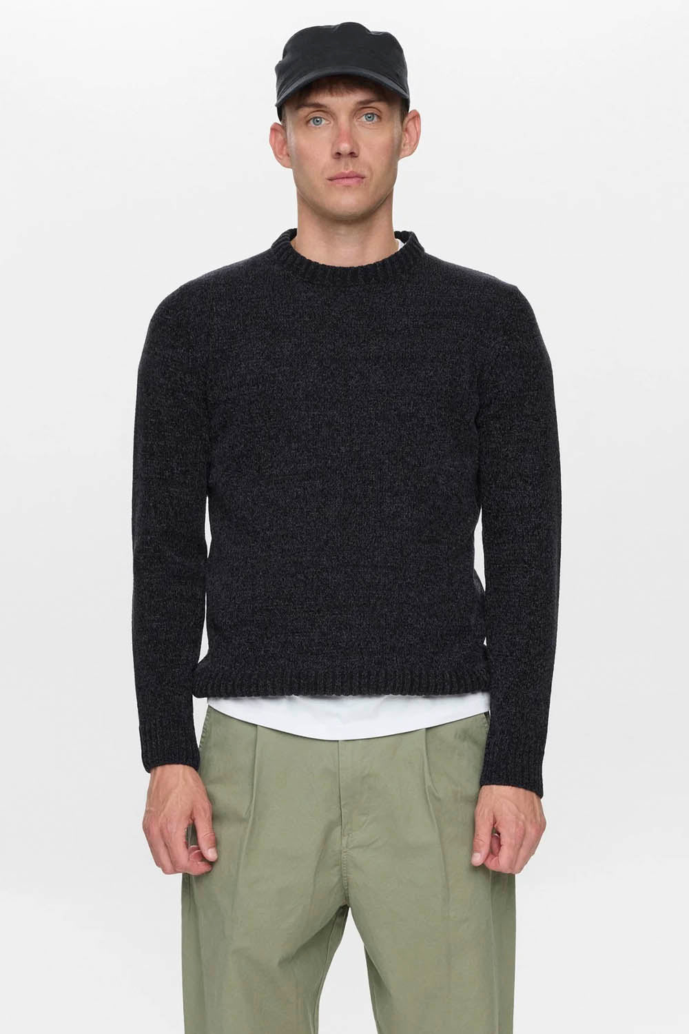 A man wearing a dark charcoal knit crewneck sweater, an olive green pair of trousers, a white undershirt, and a black baseball cap, standing against a light gray studio background.