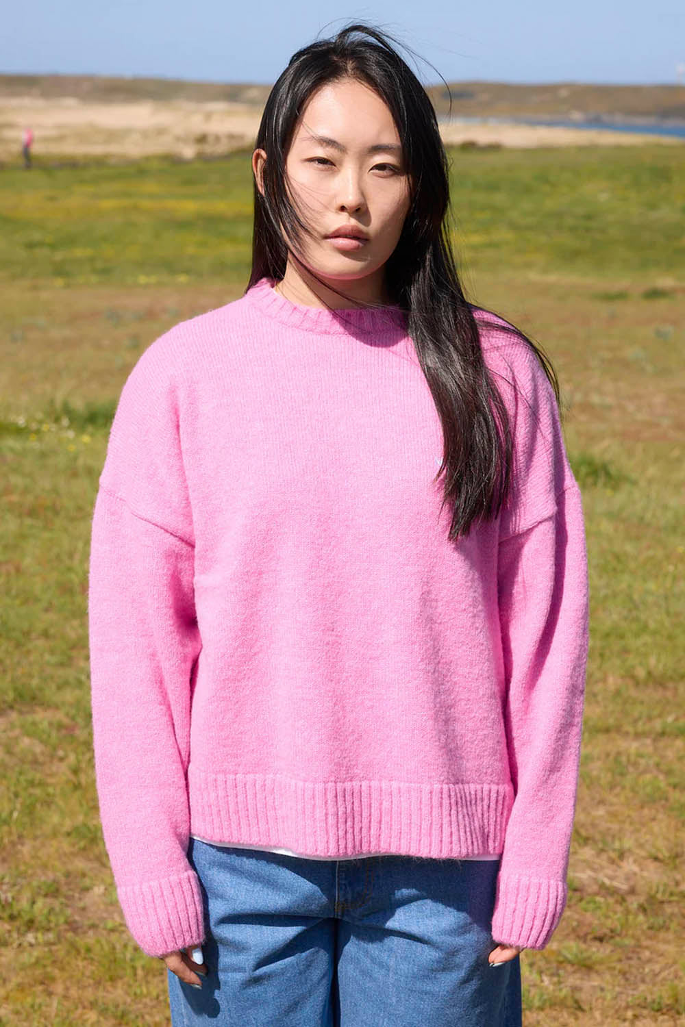 Person wearing a pink sweater and blue jeans standing in a grassy field.
