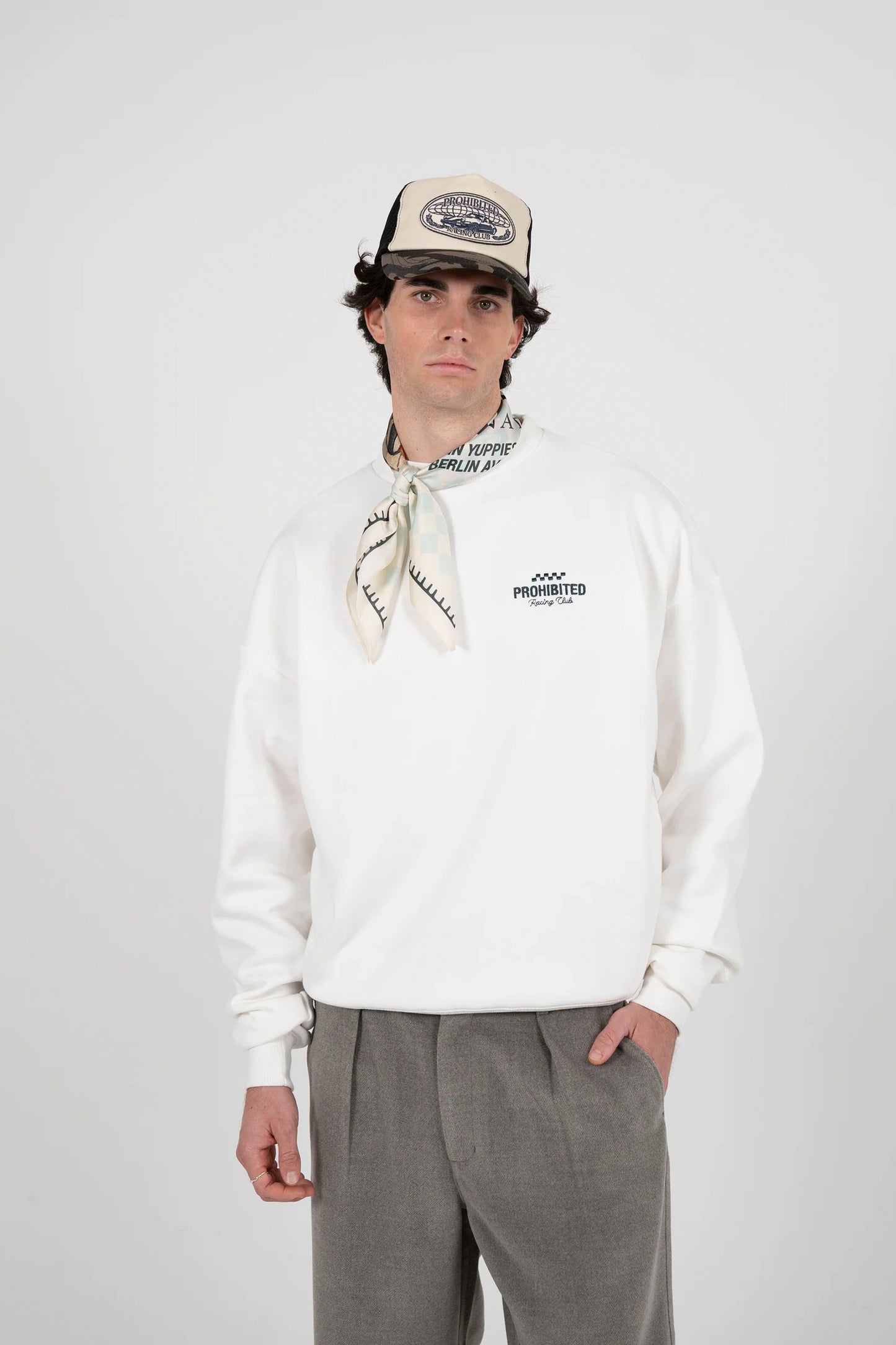 Person wearing a white PROHIBITED in a rush sweatshirt, gray pants, and a cap with a scarf on a plain background