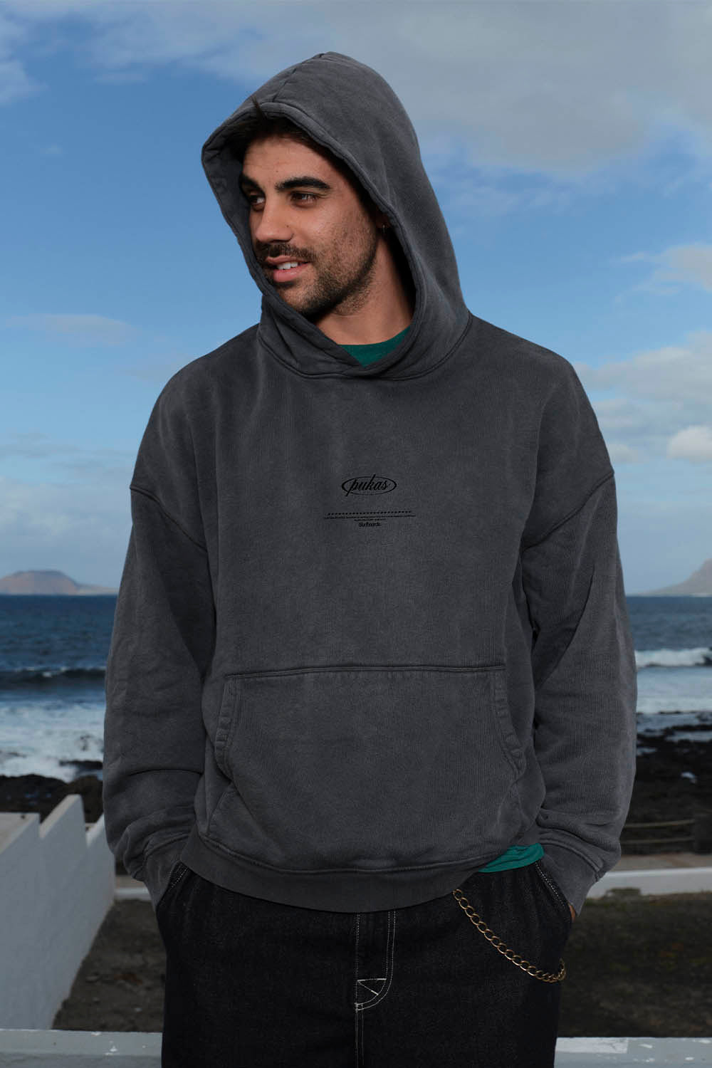 Man wearing a dark gray hoodie with a logo, standing by a coastal area with ocean and sky in the background.