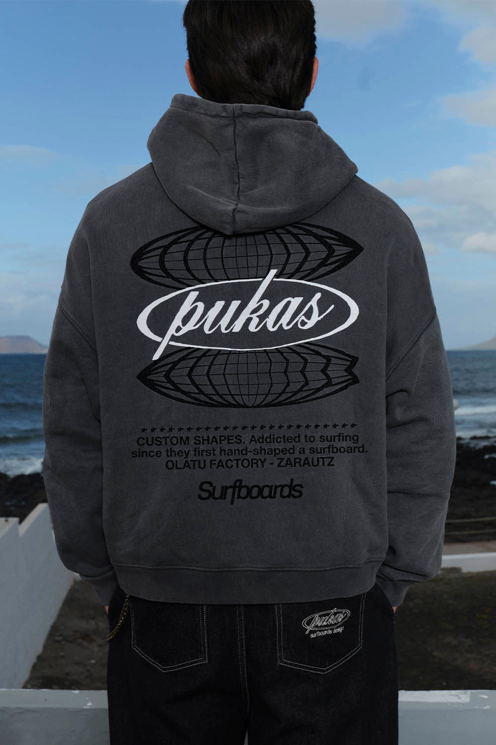 Man wearing a dark gray hoodie with a logo, standing by a coastal area with ocean and sky in the background.