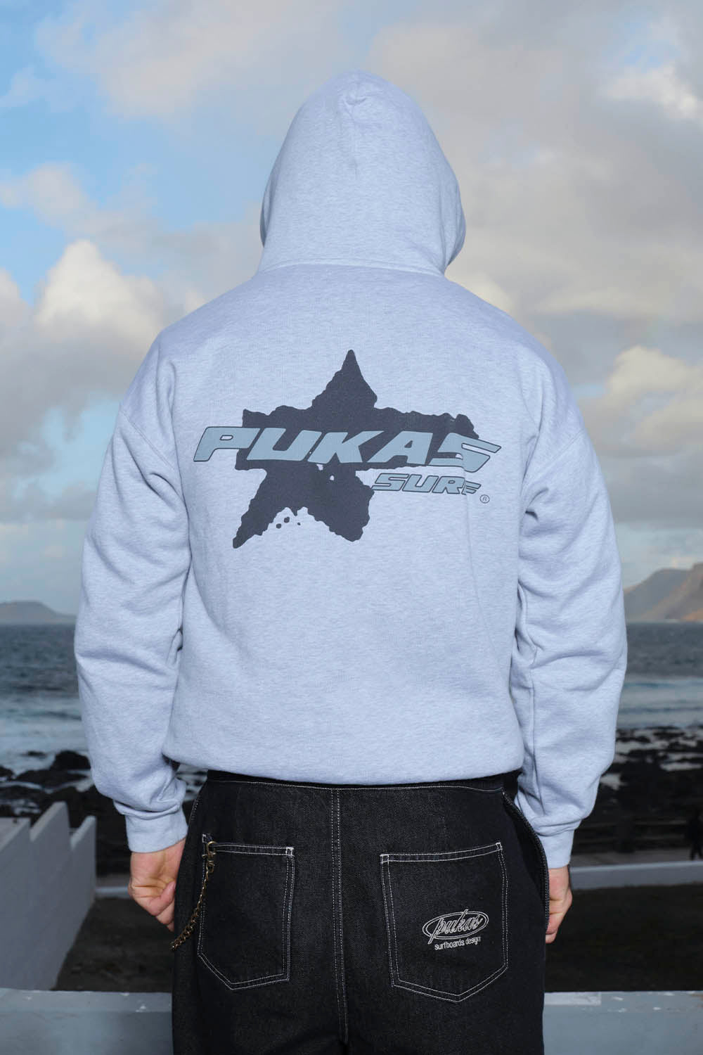 Person wearing a gray hoodie with 'Pukas Surf' logo, standing against a cloudy sky.