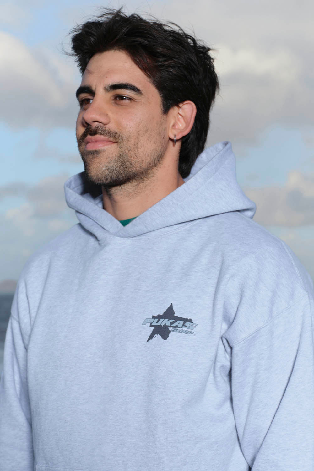 Man wearing a light gray hoodie with a logo against a cloudy sky.