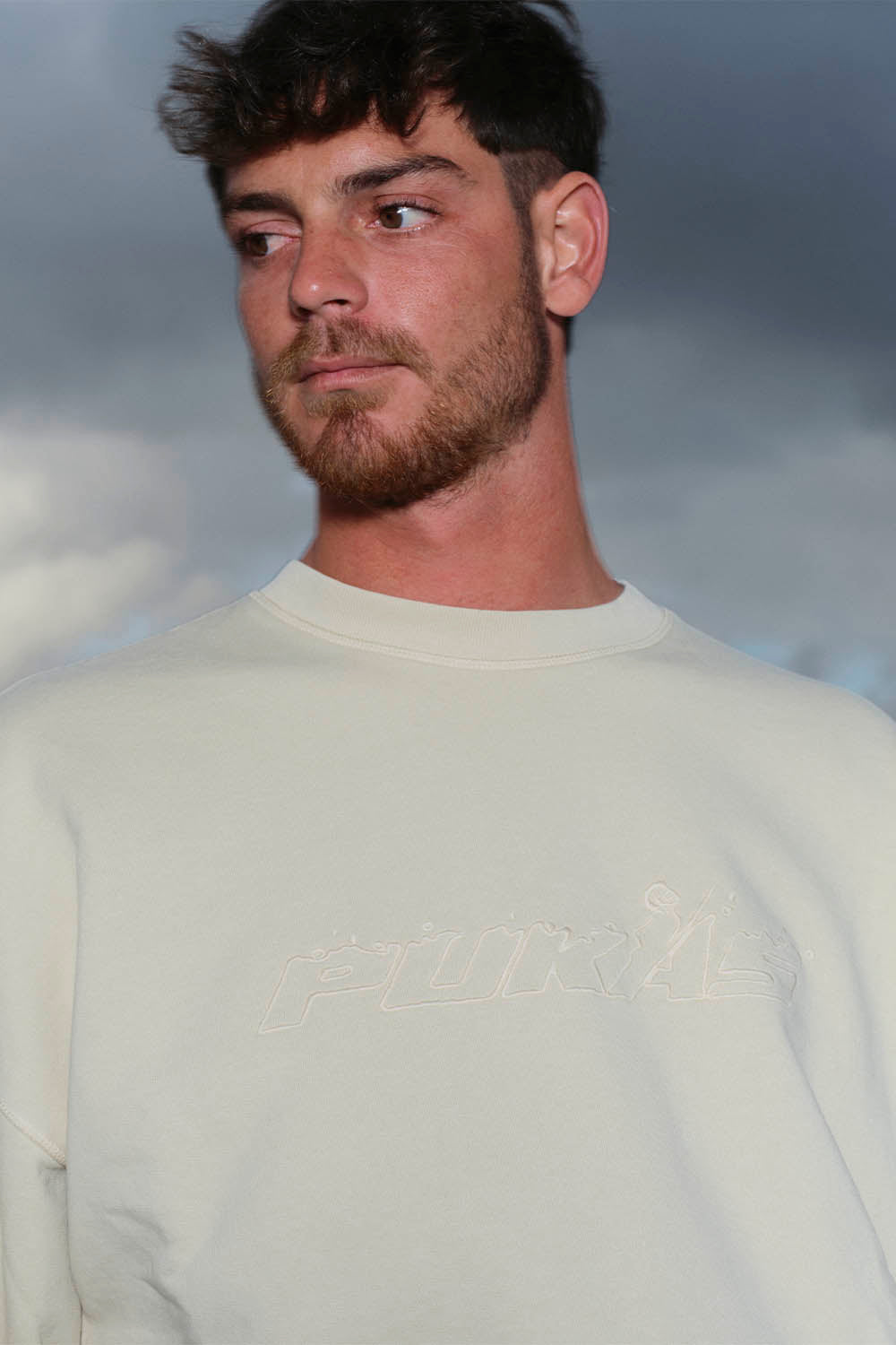 Man wearing a light-colored sweatshirt with text against a cloudy sky background