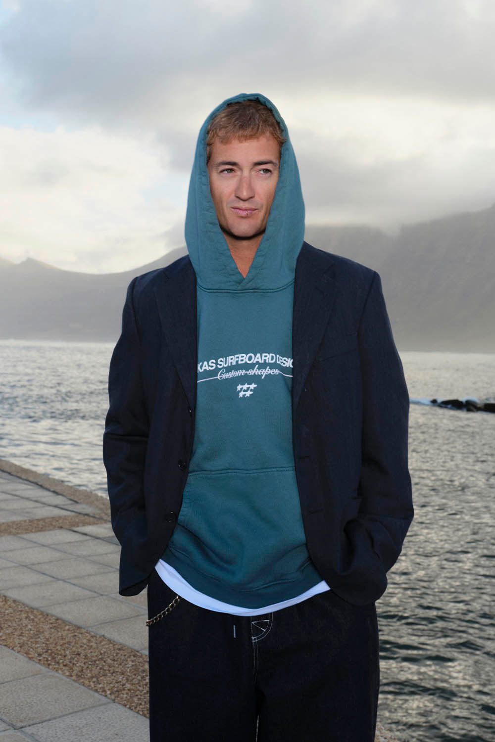 Man wearing a teal hoodie with text standing by a body of water.